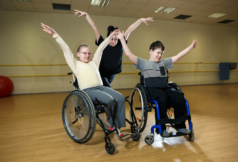 Two children in wheelchairs and an adult dance instructor raising their arms in a dance studio.